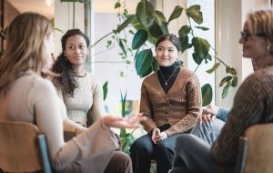 Woman sharing her experiences during a mental health group therapy meeting. Multiracial women participate in support group session sitting in a circle.